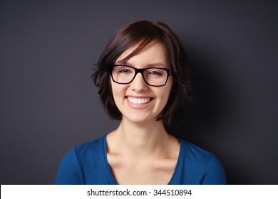 Close Up Happy Young Woman, Wearing Eyeglasses, Showing Toothy Smile At The Camera Against Gray Wall Background.