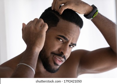 Close up head shot anxious stressed dissatisfied African American man touching head, checking hair, standing in bathroom, worried about dandruff or hair loss, healthcare and beauty concept