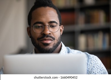 Close up head shot focused young african ethnicity man in glasses looking at laptop screen, involved in project work. Concentrated skilled millennial biracial man analyzing marketing statistics.