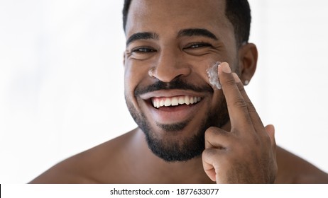 Close up head shot happy laughing young 30s african american handsome man applying moisturizing cream on cheeks, feeling excited of using professional cosmetics in domestic skincare procedure.