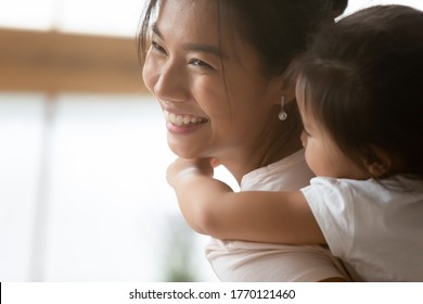 Close Up Head Shot Happy Young Asian Ethnicity Woman Carrying On Back Cute Little Baby Daughter, Smiling Biracial Female Nanny Playing With Adorable Small Kid Girl Indoors, Daycare Custody Concept.