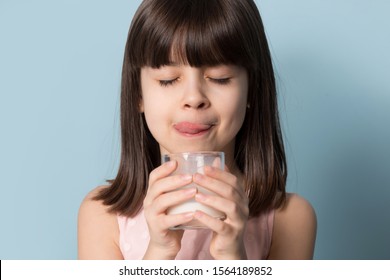 Close Up Head Shot Image Of Happy Smiling Small Girl Enjoying Glass Of Fresh Cow Milk Licking Lips. Six Year Old Child Drinking Tasty Beverage With Eyes Closed, Blue Background. Diet Nutrition Concept