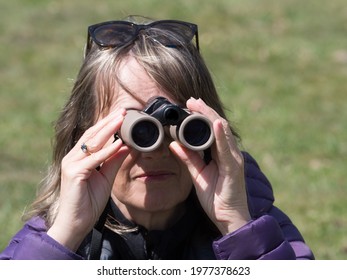 A Close Up Head Shot Of A Lady Looking Through Binoculars.She Has Sunglasses On Her Head