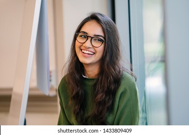 A Close Up Head Shot Portrait Of A Preppy, Young, Beautiful, Confident And Attractive Indian Asian Woman In A Green Sweater And Spectacles In A Classroom Or Office. She Is Smiling Happily. 