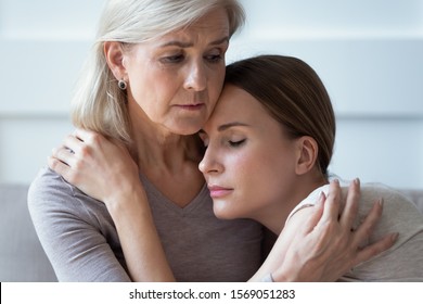 Close up head shot sad mature grey haired mother embracing, comforting upset adult daughter, sitting together on sofa. Unhappy young woman supporting stressed middle aged mommy, experiencing grief.