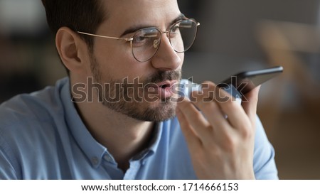 Close up head shot satisfied man wearing glasses holding smartphone near mouth, recording voice message, confident young male chatting online or speakerphone, activating digital assistant