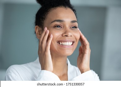 Close up head shot smiling African American young woman touching face, beautiful girl wearing white bathrobe doing facial massage, enjoying perfect smooth skin, skincare and treatment concept