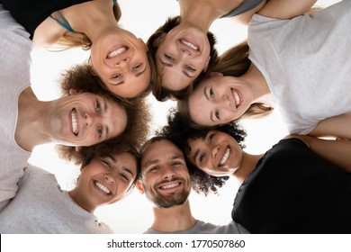 Close Up Head Shot Smiling Beautiful Mixed Race People Touching Heads In Circle, Looking Down At Camera, Healthy Multiracial Young Teammates In Sportwear Feeling Excited, Waiting For Training Workout.