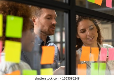 Close Up Head Shot Smiling Female African American And Caucasian Colleagues Showing Working Progress To Confident Male Team Leader On Kanban Agile Glass Window Board With Colorful Paper Stickers.