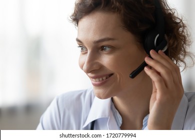 Close up head shot of smiling female young doctor operator wearing headset with microphone looking away. Medical call center healthcare services, telehealth hotline, remote tele medicine concept.
