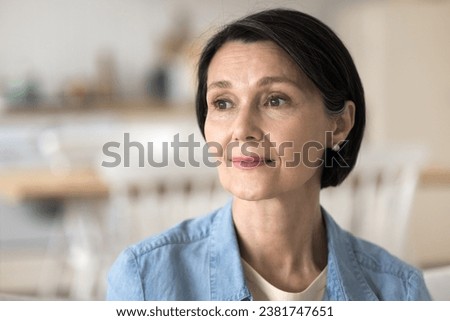 Close up head shot thoughtful middle-aged woman staring aside, deep in thoughts, having pensive mood and attractive appearance sit on sofa at home, enjoy calm retired life, remember past looks away
