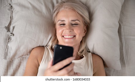 Close Up Head Shot Top Above View Happy Middle Aged Lady Lying In Bed, Looking At Smartphone Screen. Smiling Older Lady Reading Pleasant Sms, Chatting With Friends, Enjoying Lazy Weekend Morning Ime.