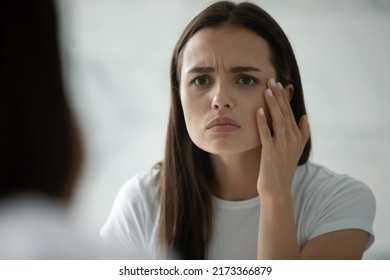 Close up head shot unhappy young woman wearing white t-shirt checking skin, looking in mirror, standing in bathroom, worried about mimic wrinkle or acne, touching face, skincare and treatment - Powered by Shutterstock