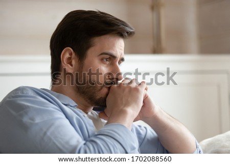 Close up head shot young thoughtful man looking away. Pensive millennial bearded guy thinking of problems, feeling stressed, sitting alone on couch at home, worrying about personal troubles.