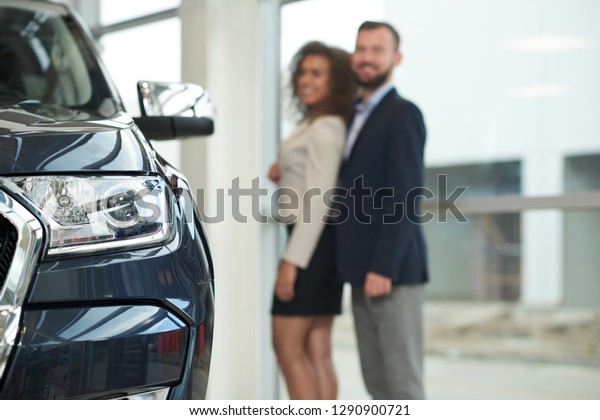 Close up of\
headlight of dark blue car. Happy couple standing behind, posing,\
looking at camera. Woman and man looking for expensive automobile\
in modern car dealership.