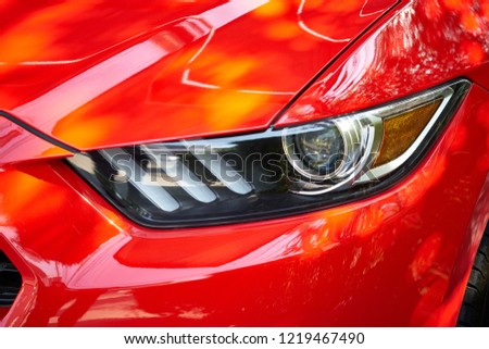 Close up of headlight detail of modern luxury sportscar with reflection on red paint after wash & wax. Front view of musclecar with brick wall. Concept of car detailing and paint protection background