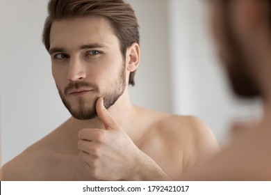 Close up headshot mirror reflection satisfied handsome young man touching beard, standing in bathroom, applying aftershave lotion, enjoying skincare procedure, morning routine concept
