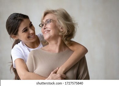 Close Up Headshot Portrait Picture Of Adult Daughter Hugs From Behind Happy Mature Mother Enjoying Tender Moment Looking At Each Other. Smiling Woman And Older Mum Embracing Having Fun Together.