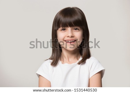 Close up headshot portrait of smiling little girl isolated on grey studio background look at camera, happy small preschooler child in white t-shirt posing feel overjoyed excited show healthy teeth