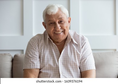Close up headshot portrait of smiling mature man sit on couch at home look at camera posing for picture, happy positive senior male or optimistic grandfather feel good relax on comfortable sofa