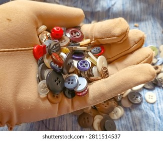 A close up high angle shot of a human hand wearing a smooth beige or tan fabric glove holding a vibrant assortment of mixed sewing buttons  The colorful collection includes buttons of various sizes shapes and colors such as red blue purple yellow black white and brown scattered across the gloved palm  The background features a soft focused textured light blue fabric surface with more scattered buttons  Bright natural lighting highlights the smooth surfaces of the plastic and metal buttons creating gentle reflections  This image is ideal for concepts of handcrafts textile work tailoring hobby crafts fashion design and sorting materials