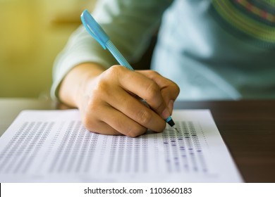 Close Up Of High School Or University Student Holding A Pen Writing On Answer Sheet Paper In Examination Room. College Students Answering Multiple Choice Questions Test In Testing Room In University.