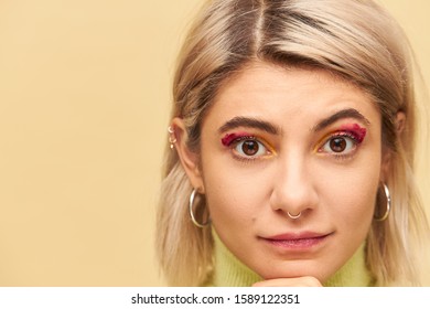Close up highly detailed image of beautiful blonde young woman wearing bob hairstyle, nose ring, round earrings and pink eye-shadows looking at camera, having serious thoughtful look, touching chin
