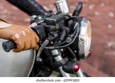 Close Up Of A Hipster Biker Man Hand In Leather Glove Hold Throttle Control Of Classic Style Cafe Racer Motorcycle. Bike Custom Made In Vintage Garage. Brutal Fun Urban Lifestyle. Outdoor Portrait.