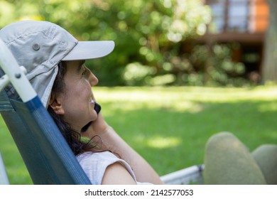 Close Up Of Hispanic Young Woman Wearing A Cap Talking With On Mobile Phone And Doing Home Office While Sitting In A Lounge Chair At The Backyard In The Springtime. Copy Space.