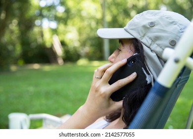 Close Up Of Hispanic Young Woman Wearing A Cap Talking With On Mobile Phone And Doing Home Office While Sitting In A Lounge Chair At The Backyard In The Springtime. Copy Space.