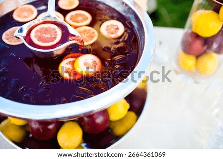 Close up horizontal image of a red punch with yellow lemons on a hot summer day being poured out of a punch bowl with a silver ladle. 