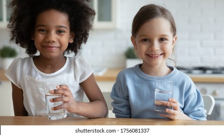 Close Up Horizontal Image Two School Age Girls African Caucasian Little Adorable Kids Sit In Kitchen Holding Glasses With Still Clear Water Look At Camera. Healthy Lifestyle, Natural Hydration Concept