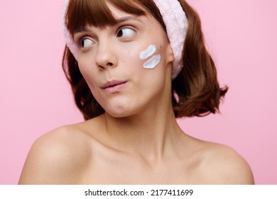 a close horizontal portrait on a pink background, of a funny, attractive woman with cream on her face, and a soft pink rim on her head, looks funny to the side