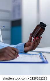 Close Up Of Hospital General Practitioner Specialist Holding A Vial Of Medicine While Checking Symptom List And Patient Illness History. Medic Having Antiviral Bottle In Hand
