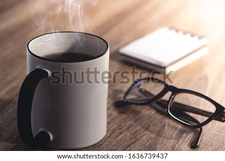 Close up of a hot black and white steaming cup of coffee/tea, modern black glasses and a note book on a wooden desk with intense sun light coming from the side. Relaxation time for drink background.