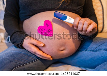 A close up hot on the large stomach of a heavily pregnant woman with a pink heart saying its a girl, she holds positive pregnancy test stick in hand