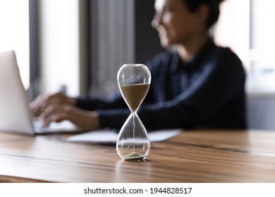 Close up hourglass measuring time, standing on wooden office table, Indian businesswoman working on background, efficiency, deadline and time management concept, busy employee using laptop - Shutterstock ID 1944828517