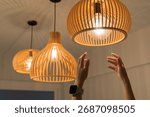 Close up of human hands reaching to adjust glowing wooden pendant ceiling lamp. Modern home interior decoration, warm lighting design, stylish contemporary atmosphere for living space, or restaurant.