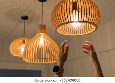 Close up of human hands reaching to adjust glowing wooden pendant ceiling lamp. Modern home interior decoration, warm lighting design, stylish contemporary atmosphere for living space, or restaurant.