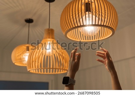 Close up of human hands reaching to adjust glowing wooden pendant ceiling lamp. Modern home interior decoration, warm lighting design, stylish contemporary atmosphere for living space, or restaurant.