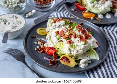 Close up of ice berg lettuce wedge salad with blue cheese dressing, bacon, tomatoes, onions, chives sitting on gray stoneware plates. Surrounded by ingredients. Keto.