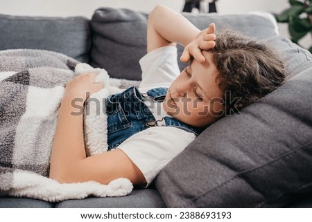 Close up of ill unhealthy little girl child lying alone on couch under blanket in living room at home. Upset small caucasian kid has fever. Concept of health, illness, sickness, common cold, treatment