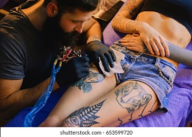 Close Up Image Of The Bearded Tattoo Male Artist Makes A Tattoo On A Female Leg.