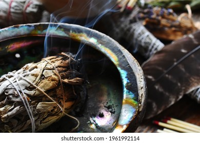 A close up image of a burning white sage smudge stick, healing crystals, and sacred feather. 