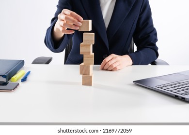 A Close Up Image Of Bussines Woman Woman Stacking Wooden Blocks In Her Work Enviroment. Office Interior Laptop, Glass Of Water And Notebook On White Desk.