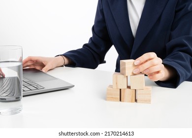 A Close Up Image Of Bussines Woman Woman Stacking Wooden Blocks In Her Work Enviroment. Office Interior Laptop, Glass Of Water And Notebook On White Desk.