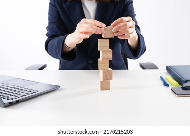 A Close Up Image Of Bussines Woman Woman Stacking Wooden Blocks In Her Work Enviroment. Office Interior Laptop, Glass Of Water And Notebook On White Desk.