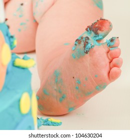 close up image of a Caucasian baby boys,dirty blue icing and chocolate cake covered leg,foot and toes, with his blue,yellow and orange polka doted birthday cake on a cream backdrop.