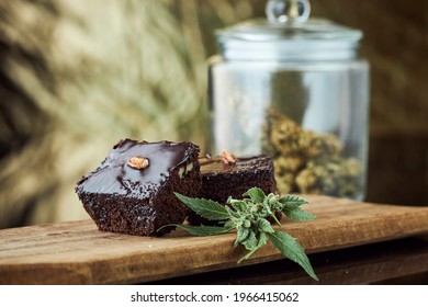 A close up image of a chocolate brownies infused with marijuana with focus on a fresh marijuana head in studio still life with glass jar of marijuana nuggets in background