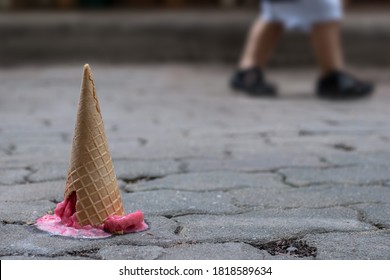 Close up image of fallen pink strawberry sorbet ice cream with cracking cone on the old cement ground and blurred children legs background. Selective focus and copy space. 